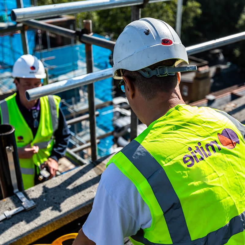 Two Mitie workers on scaffolding, wearing high vis vests and hard hats